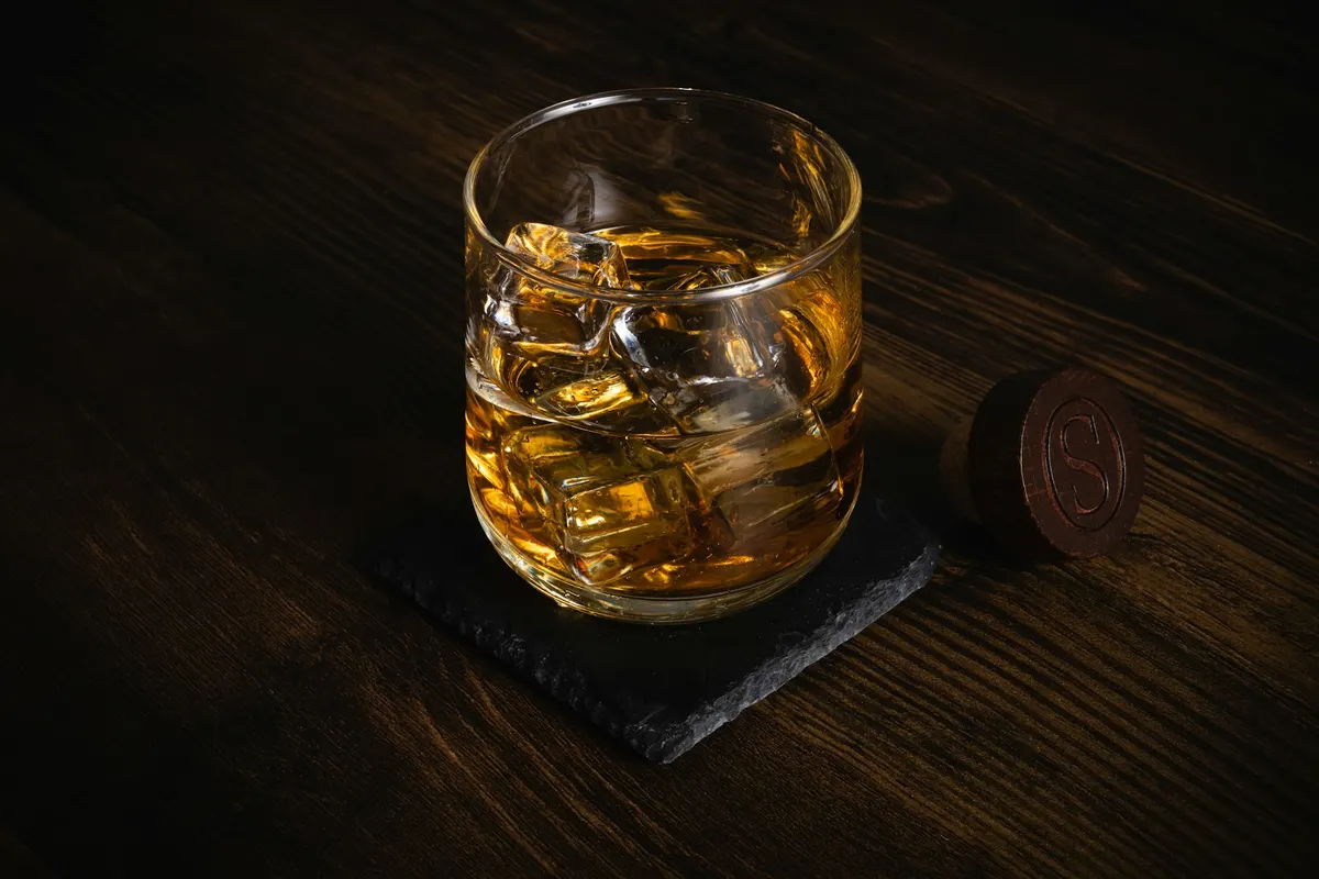 A glass of whiskey with ice on a slate coaster on a dark wooden table